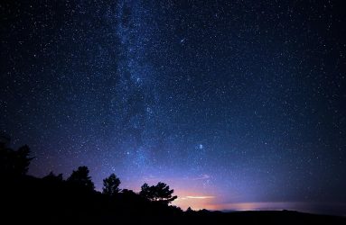 photo of silhouette of trees on hillside with backdrop of starry deep blue sky and faint golden sunlight on horizon