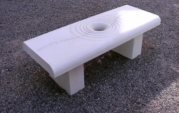 Carved stone bench with hole in centre and concentric circles like ripples, line of poetry